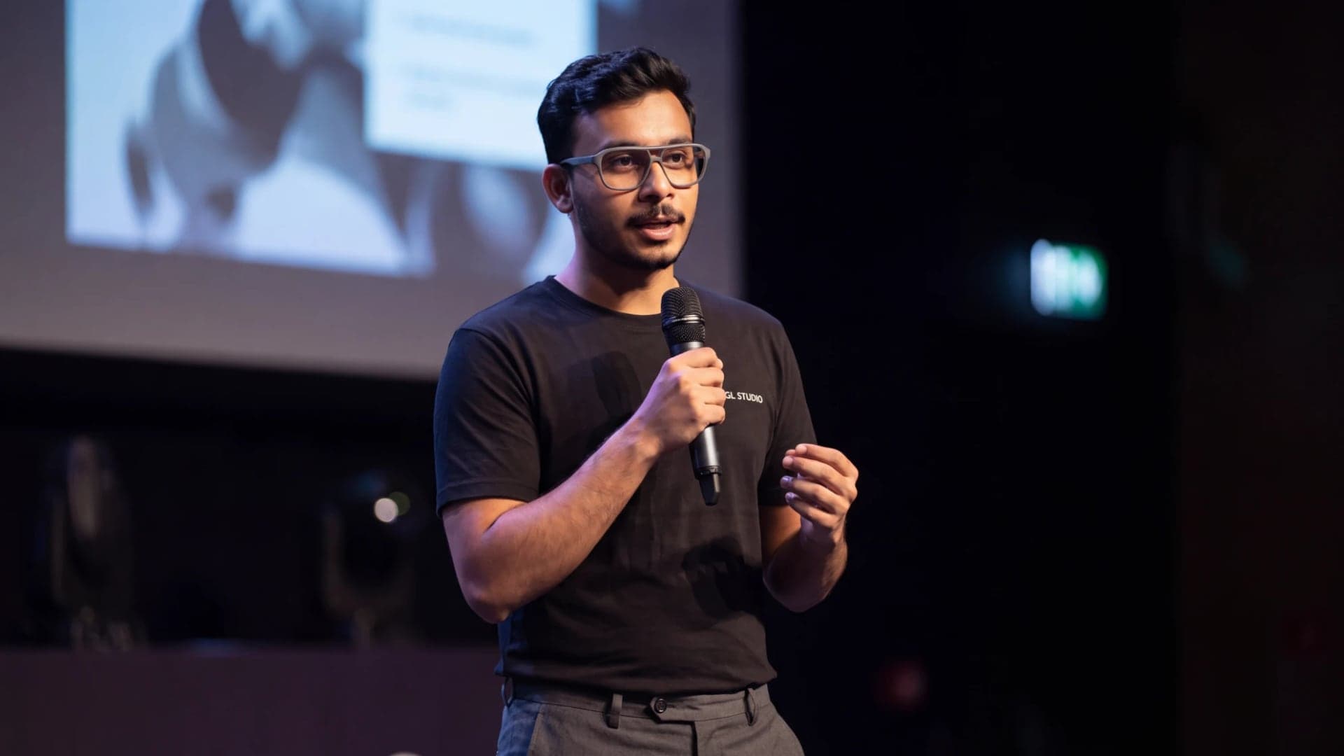 Kamran Ali speaking on stage into a handheld microphone, wearing a black Jugl Studio T-shirt, a projected slide behind him.